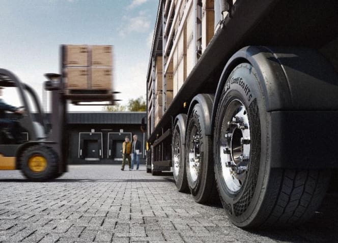 Conti Eco Ht 51 | ยางรถยนต์ ราคาอัปเดต 2026 ครบทุกยี่ห้อ รวมยาง Ev | Autotirechecking Forklift lifting a pallet beside a large trailer in a loading yard; two workers stand nearby.