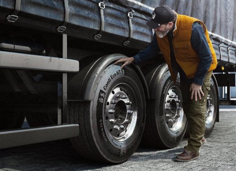 Man in an orange safety vest inspecting large trailer tires beside a parked truck.