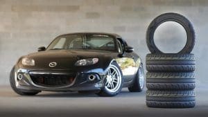 Black Mazda MX-5 Miata parked in a garage beside a vertical stack of four tires, with one tire upright next to it.
