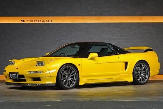 Bright yellow two-door sports car with a black roof and large rear spoiler, parked indoors in a showroom wall setting.