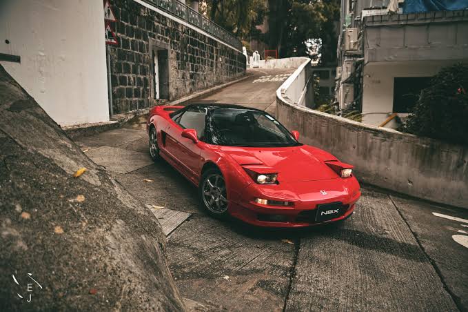 Red Acura NSX sports car on a winding city street, curving guardrail and stone wall in the background.