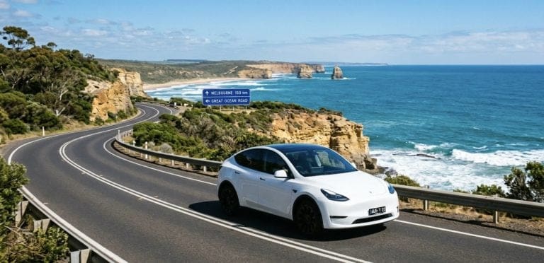 White electric car on a winding coastal road with rocky cliffs and the blue ocean; road signs indicate Melbourne 150 km and the Great Ocean Road.