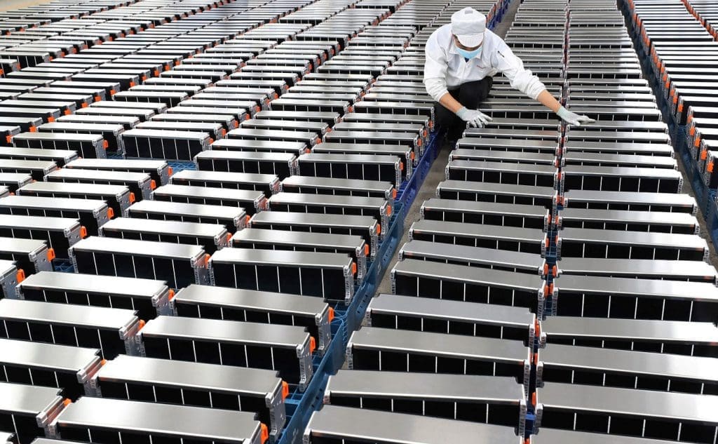 Img3394 | ยางรถยนต์ ราคาอัปเดต 2026 ครบทุกยี่ห้อ รวมยาง Ev | Autotirechecking Worker in a white cleanroom suit, mask, and gloves inspecting rows of long battery modules arranged on racks in a manufacturing facility.