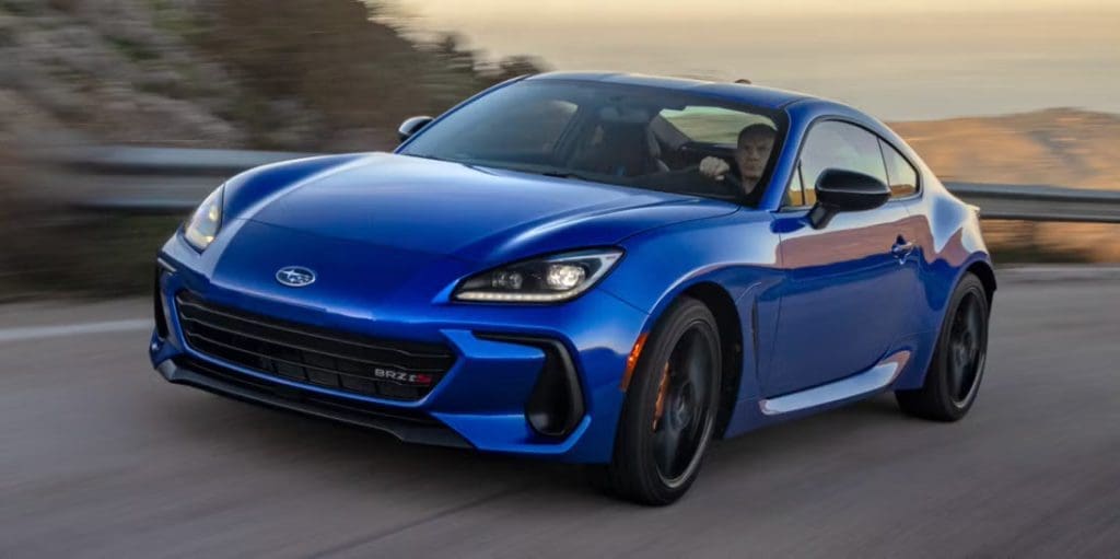 Img4023 | ยางรถยนต์ ราคาอัปเดต 2026 ครบทุกยี่ห้อ รวมยาง Ev | Autotirechecking Blue sports car in motion on a curving road, driver visible through the window with motion blur in the background.