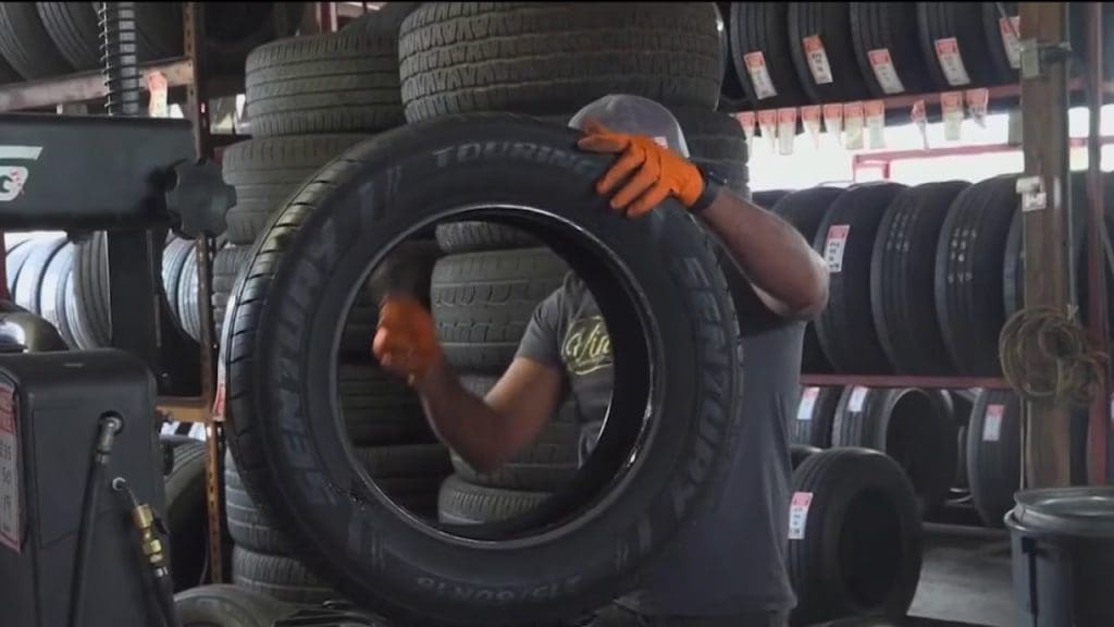 Img4212 | ยางรถยนต์ ราคาอัปเดต 2026 ครบทุกยี่ห้อ รวมยาง Ev | Autotirechecking Worker wearing gloves holds a large tire up in a tire shop, with stacked tires on shelves in the background as other tires sit nearby.
