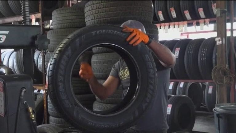 Worker wearing gloves holds a large tire up in a tire shop, with stacked tires on shelves in the background as other tires sit nearby.
