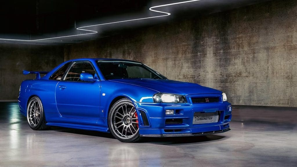 Blue Nissan Skyline GT-R-style sports car parked in a dim concrete garage with neon ceiling lights and aftermarket wheels on the front wheel.