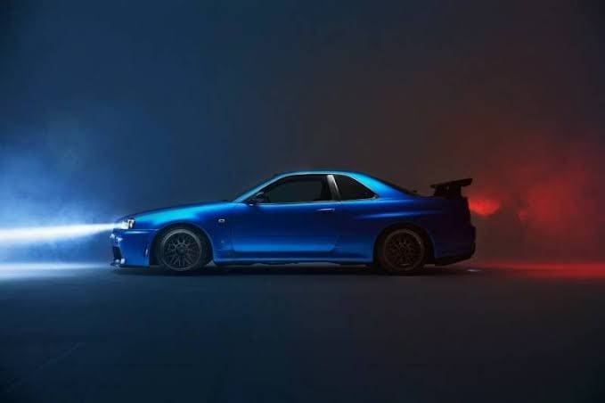 Blue sports car with a large rear wing, shown in a dark setting with blue headlights and red smoke around it.