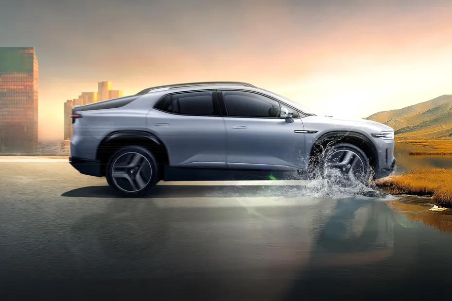 Img4448 | ยางรถยนต์ ราคาอัปเดต 2026 ครบทุกยี่ห้อ รวมยาง Ev | Autotirechecking Silver SUV drives through a puddle, water splashing, with city skyline to the left and hills to the right at sunset in the background.