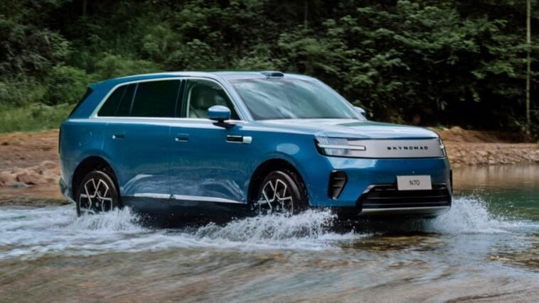 Blue electric SUV crossing a shallow river in a forested area, creating splashes around the wheels.