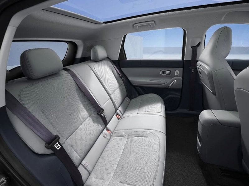 Rear passenger seats in a modern car with light gray fabric upholstery, three-seat bench and visible seat belts, sunroof overhead.