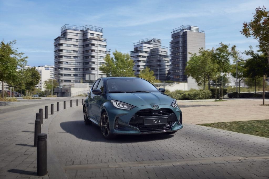Img4884 | ยางรถยนต์ ราคาอัปเดต 2026 ครบทุกยี่ห้อ รวมยาง Ev | Autotirechecking Blue Toyota Yaris driving on a curved city street with modern apartment buildings in the background.