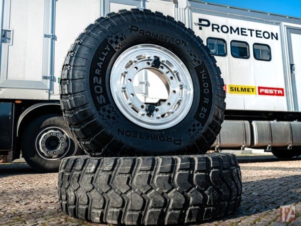 Img5059 | ยางรถยนต์ ราคาอัปเดต 2026 ครบทุกยี่ห้อ รวมยาง Ev | Autotirechecking Large off-road tire stacked on top of another tire in front of a Prometeon truck trailer; rugged industrial scene.
