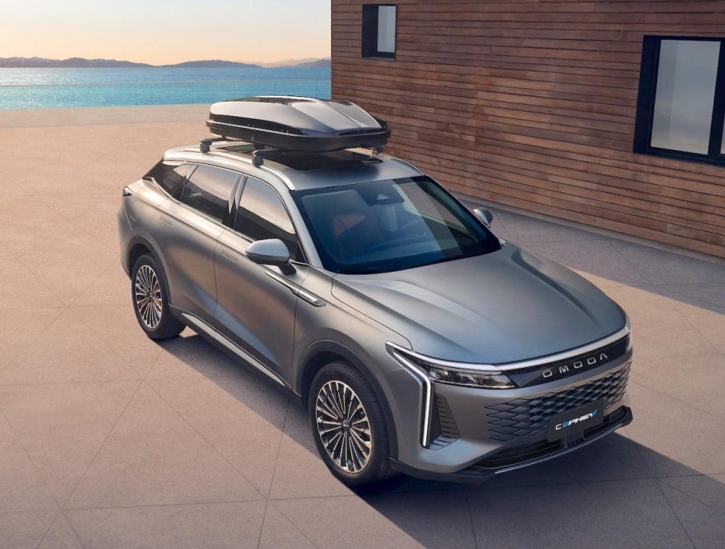 Omoda C9 Phev 20265 | ยางรถยนต์ ราคาอัปเดต 2026 ครบทุกยี่ห้อ รวมยาง Ev | Autotirechecking Silver SUV with a rooftop cargo box parked on a light concrete surface beside a wooden building and the sea in the background.