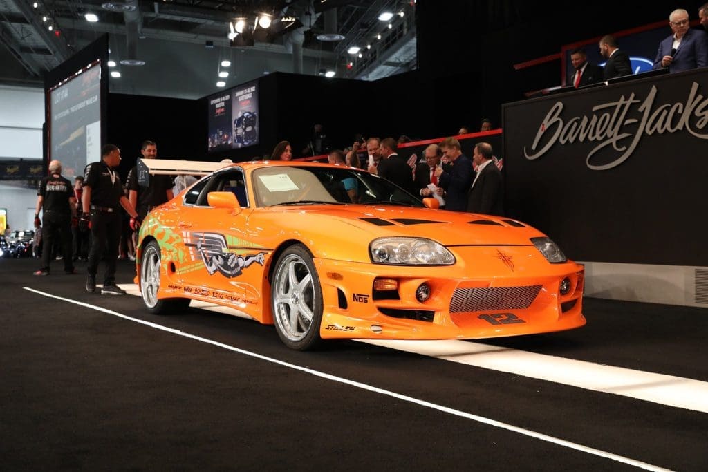 Orange sports car on a showroom floor at an auto show, with a crowd and a Barrett-Jackson banner in the background, spotlighting its sleek body and custom graphics.