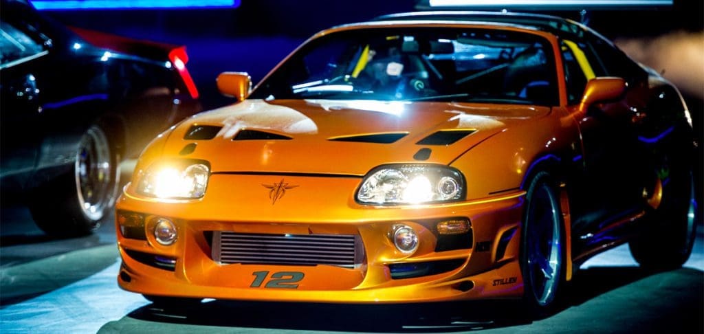 Orange sport coupe on a showroom floor with headlights on and the number 12 on the front bumper, showcasing a modified performance build at night.
