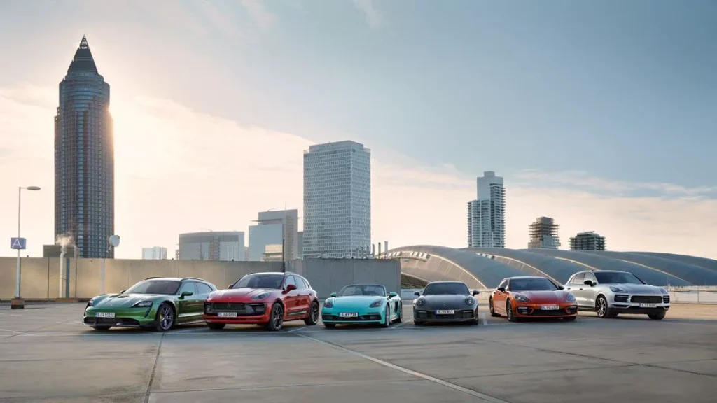 Seven colorful sports cars parked in a row on a rooftop parking area with a modern city skyline in the background at sunset/sun glare on the left.