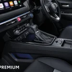 Interior of a car with a leather-wrapped center console, gear shifter, digital touchscreen on the dash, and a steering wheel; 'HEV PREMIUM' text visible in the lower left corner.